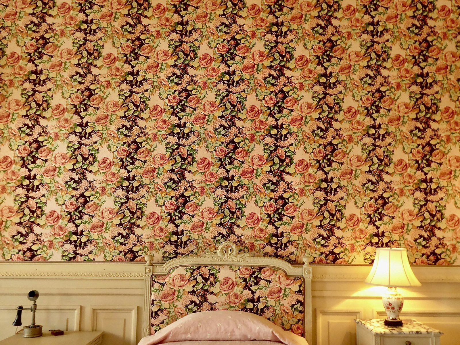 Bedroom wall covered in rose-and-cream floral wallpaper behind a cream upholstered headboard and a glowing bedside lamp
