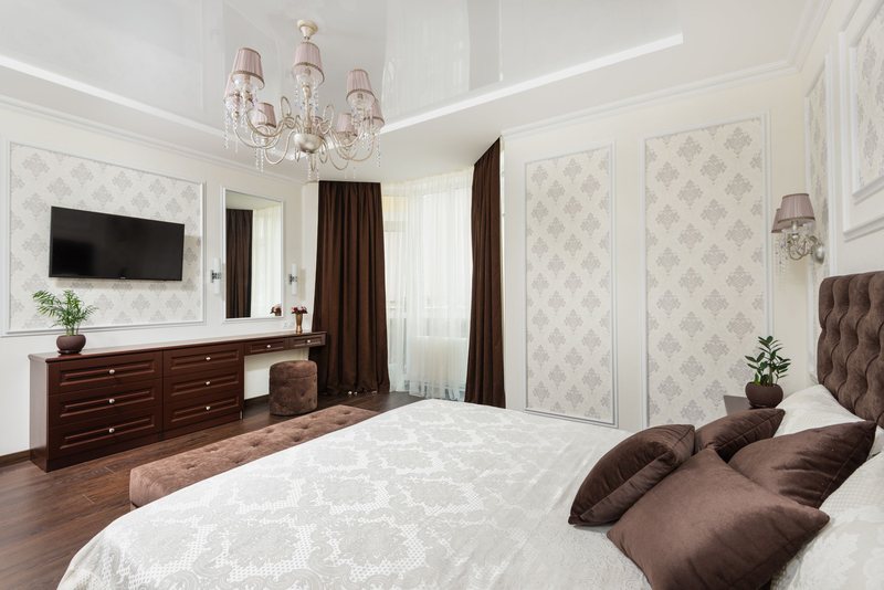 Bright bedroom with cream damask wallpaper set inside framed wall panels, a crystal chandelier and a tufted headboard