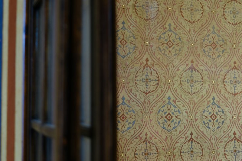 Close view of an ornate ogee-pattern wallpaper in gold, rust and blue beside a dark wooden window frame