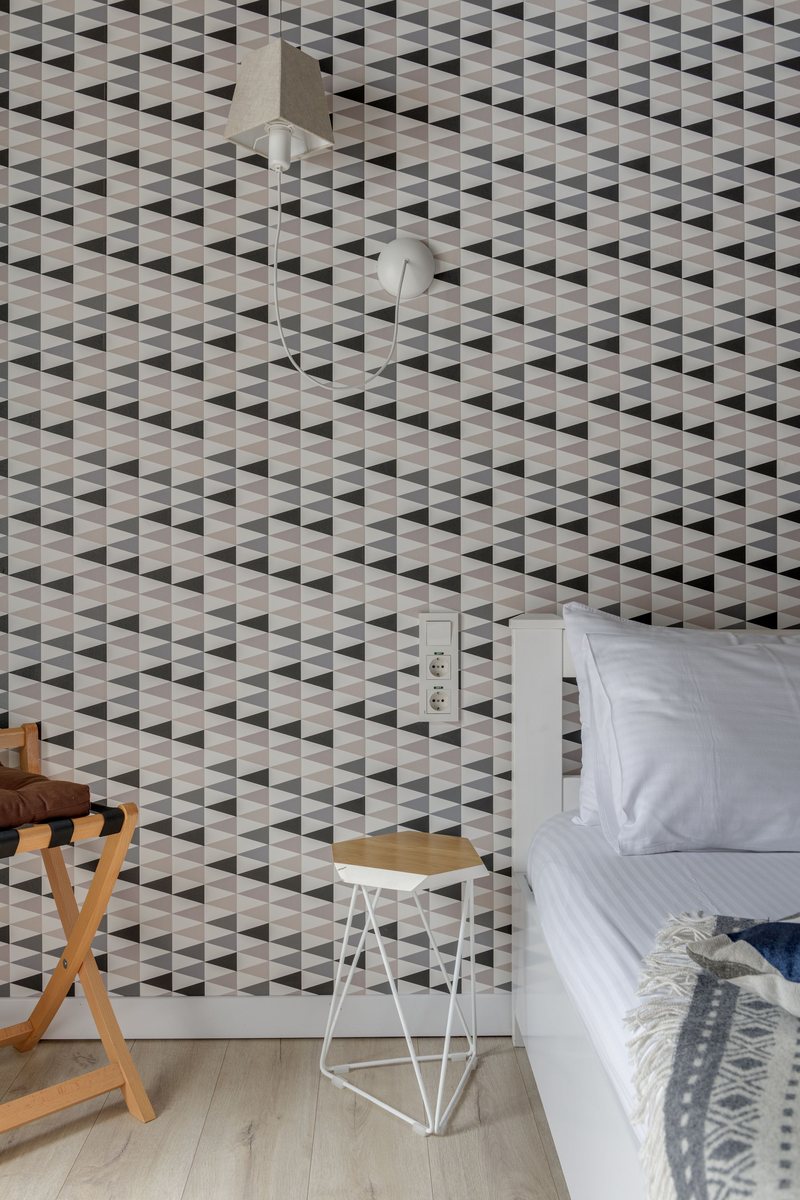 Bedroom corner with black, grey and white triangle-pattern wallpaper, a white bed and a small geometric stool