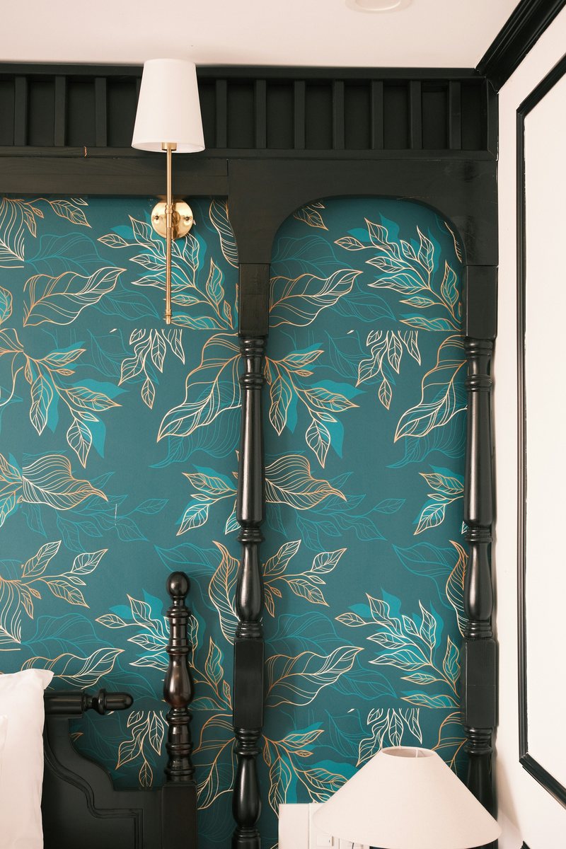 Teal wallpaper with gold-outlined leaves framed by a black four-poster bed and a brass wall sconce