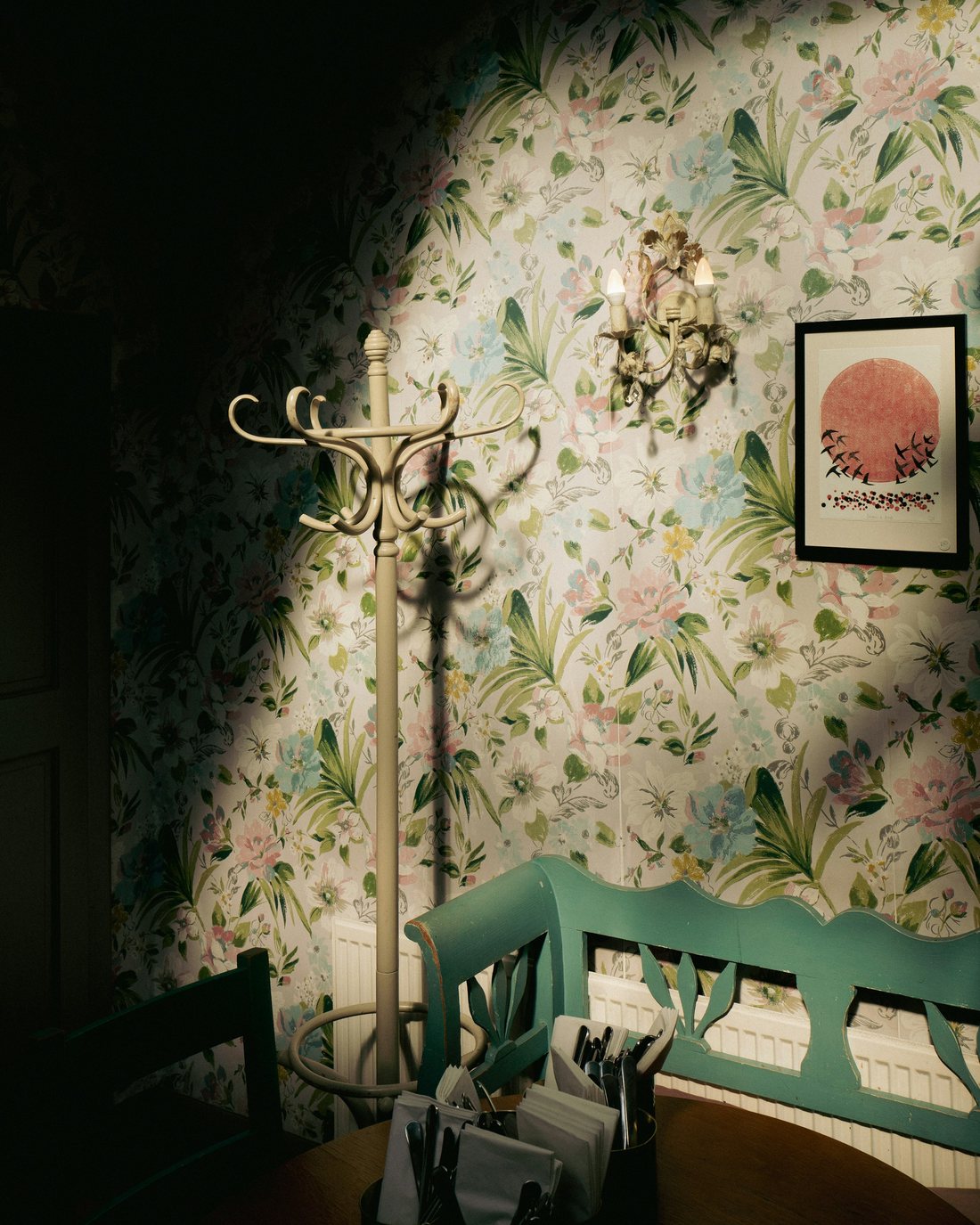 Softly lit hallway corner with pastel floral wallpaper, a brass wall sconce, a bentwood coat stand and a green painted bench