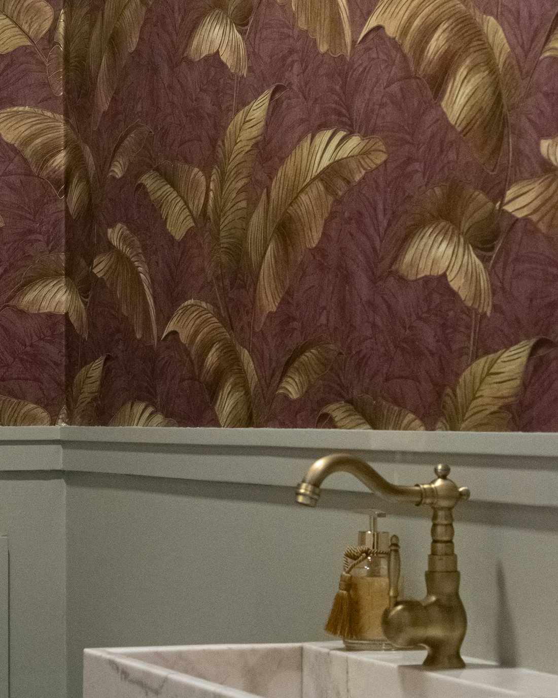 Powder room with plum wallpaper printed with large gold palm leaves above sage wainscoting, a brass faucet and a marble basin